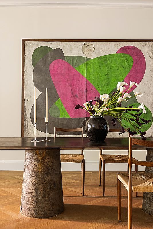 This elegant dining room features a substantial dark wood table supported by a unique, textured cylindrical pedestal base. The space is anchored by a large, abstract art piece with bold green and pink shapes, while a dark ceramic vase filled with calla lilies serves as a sophisticated centerpiece. The room is finished with warm-toned herringbone wood flooring and minimalist wooden chairs with woven seats, creating a refined and artistic atmosphere.