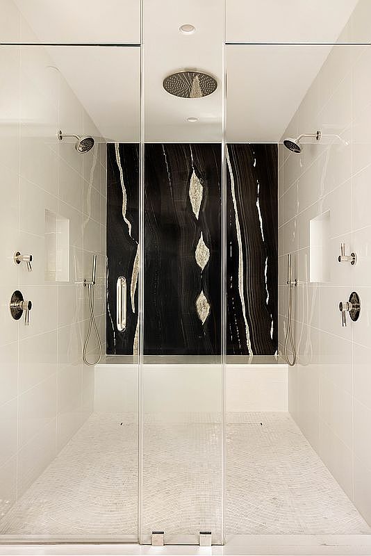 This luxurious walk-in shower features a symmetrical design with dual shower heads and handheld sprayers on either side. A striking, dark stone slab with prominent light veining serves as a dramatic focal point on the back wall, complemented by white tiled side walls and a textured, light-colored stone floor. The space is enclosed by clear glass panels, creating an open and sophisticated atmosphere.