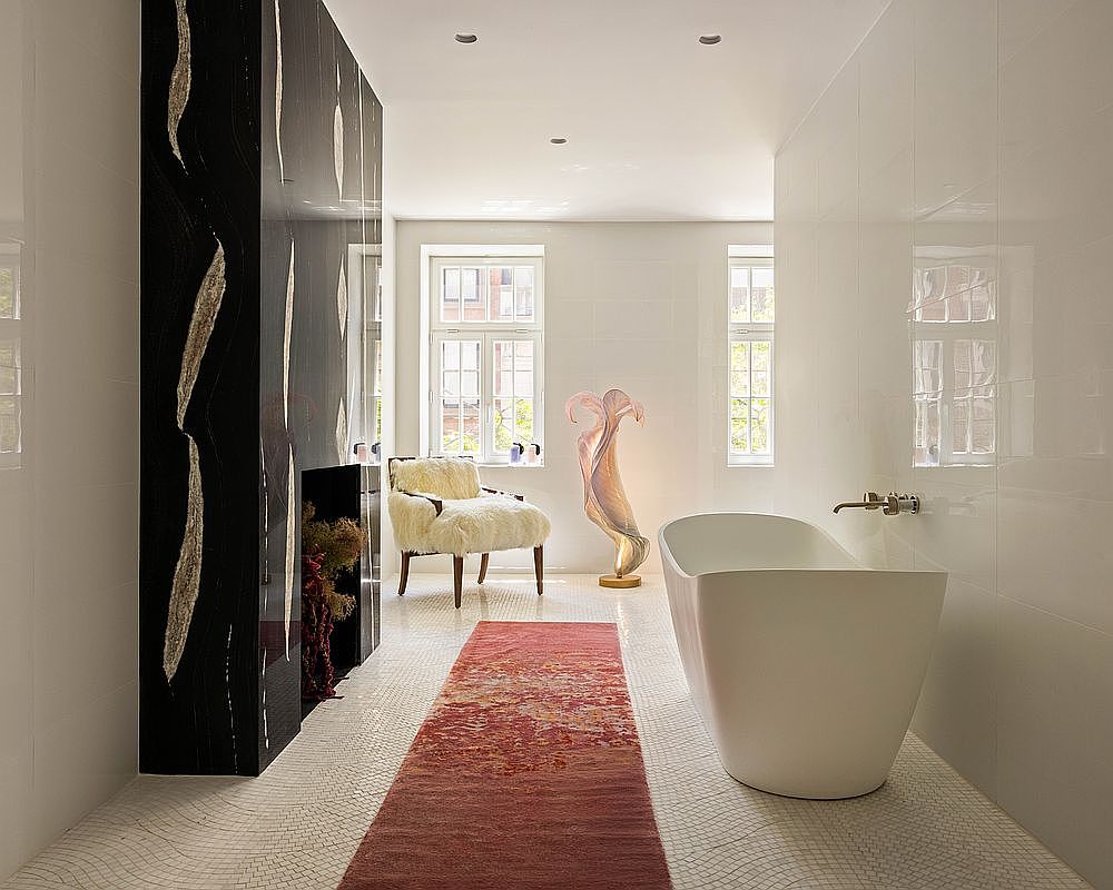 This luxurious primary bathroom features a sleek, freestanding white soaking tub positioned against a wall of glossy white tiles. To the left, a striking black wall with textured, light-colored vertical accents creates a dramatic focal point, while a plush, cream-colored armchair sits near the windows. A long, vibrant red runner rug stretches across the mosaic-tiled floor, leading the eye toward the bright, natural light streaming in from the windows.
