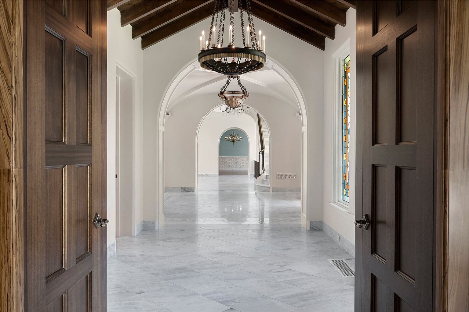 This interior shot showcases a grand hallway with a series of arched doorways leading to other rooms. The hallway features marble flooring laid in a diamond pattern, a decorative chandelier, and a glimpse of a staircase in the distance. The open doors on either side of the frame invite viewers into the space, creating a sense of depth and luxury.