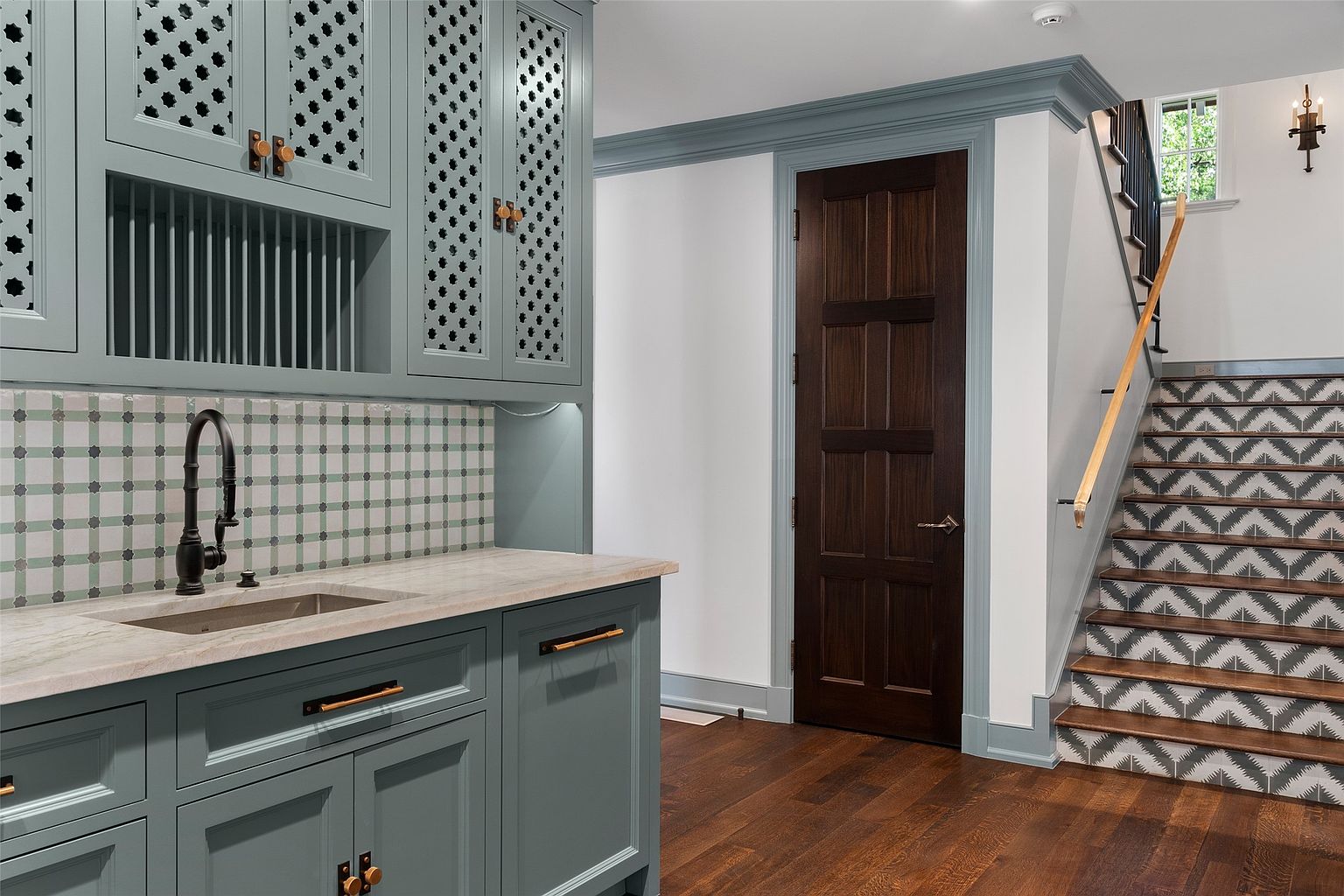 This interior shot showcases a stylish hallway with a unique staircase design. The staircase features patterned risers and a wooden handrail, complemented by a dark wood door and custom cabinetry with a sink. The color palette includes muted blues and browns, creating a sophisticated and inviting atmosphere.