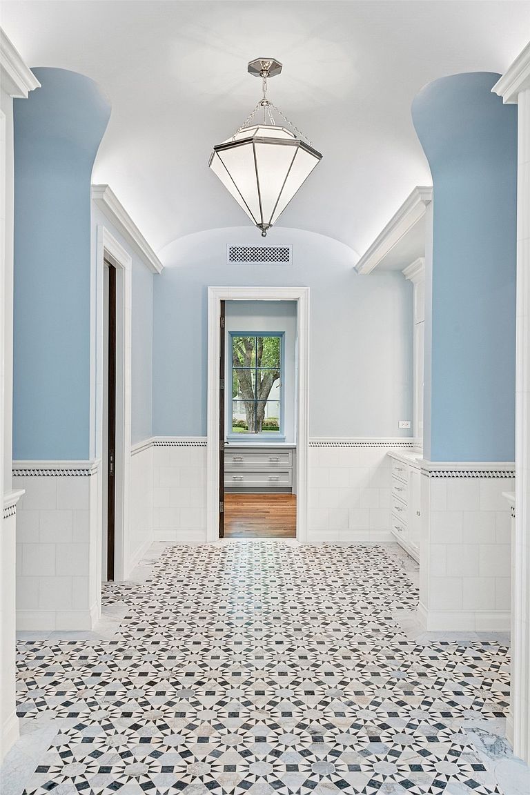 This interior shot showcases a stylish hallway with a unique geometric tile floor and light blue walls. The hallway features white wainscoting, arched entryways, and a decorative pendant light. A doorway at the end of the hall provides a glimpse into another room, adding depth and interest to the space.