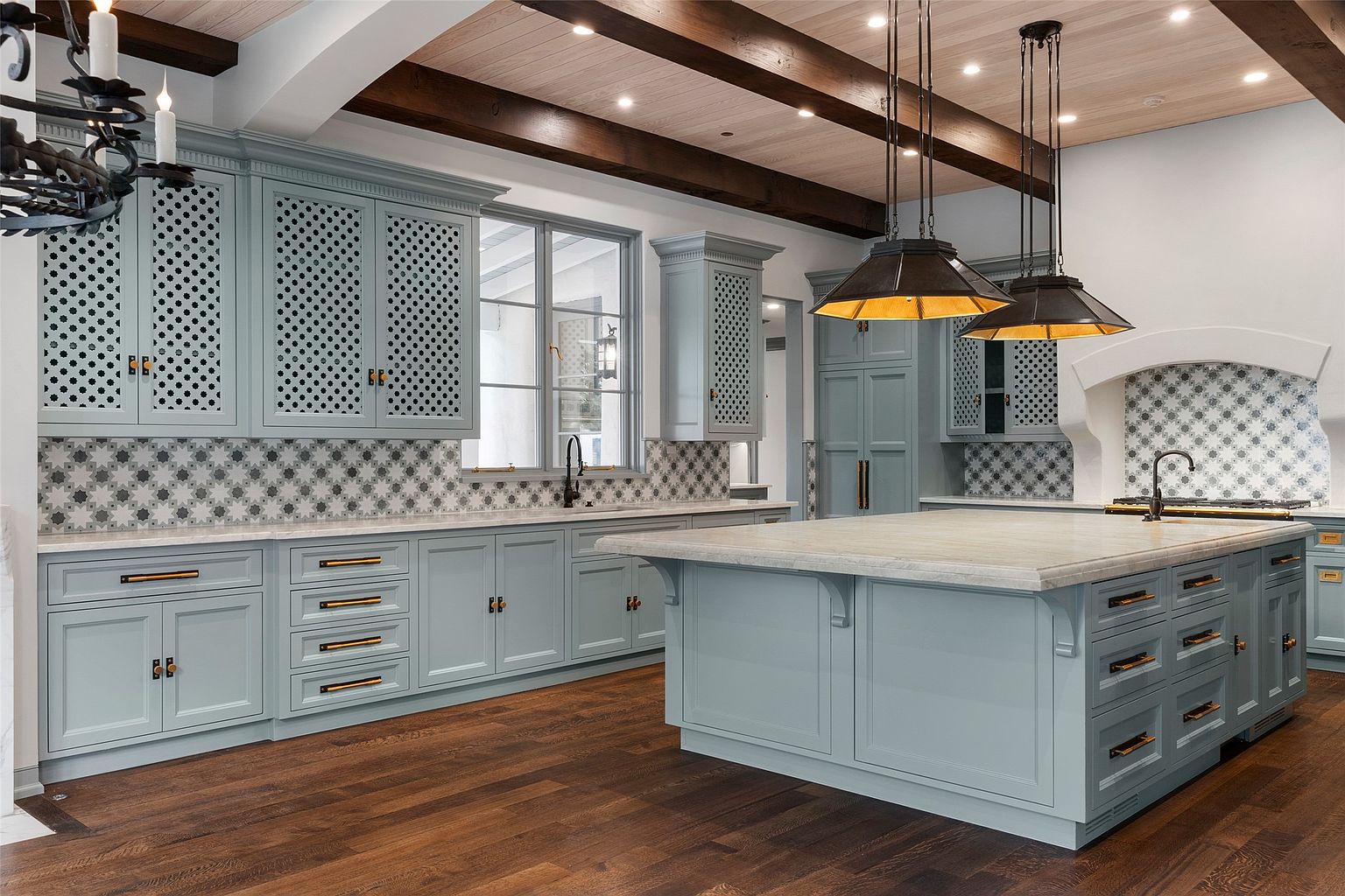 This is a well-lit kitchen featuring custom blue-grey cabinetry with decorative lattice inserts on the upper cabinets and bronze hardware. A large island with a marble countertop sits in the center of the room, complemented by dark hardwood flooring and exposed wooden beams on the ceiling. Three pendant lights hang above the island, adding a touch of modern elegance to the space.