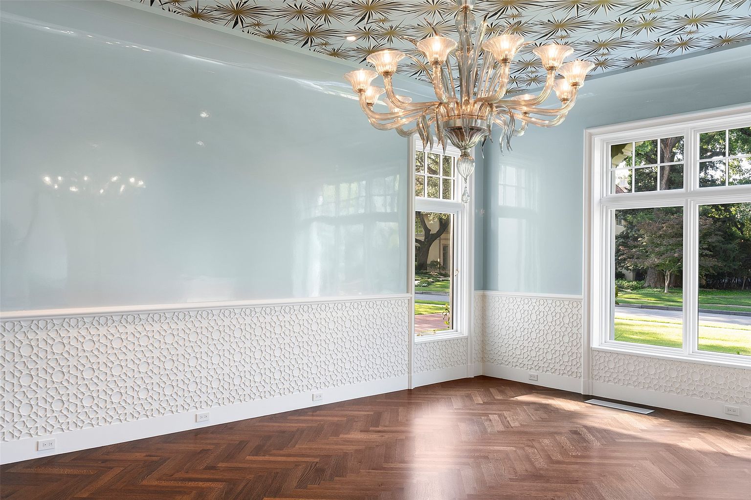 This elegant dining room features light blue walls, decorative white wainscoting, and a herringbone wood floor. A large crystal chandelier hangs from the patterned ceiling, adding a touch of luxury. Large windows provide ample natural light and views of the outdoors, creating a bright and inviting space.