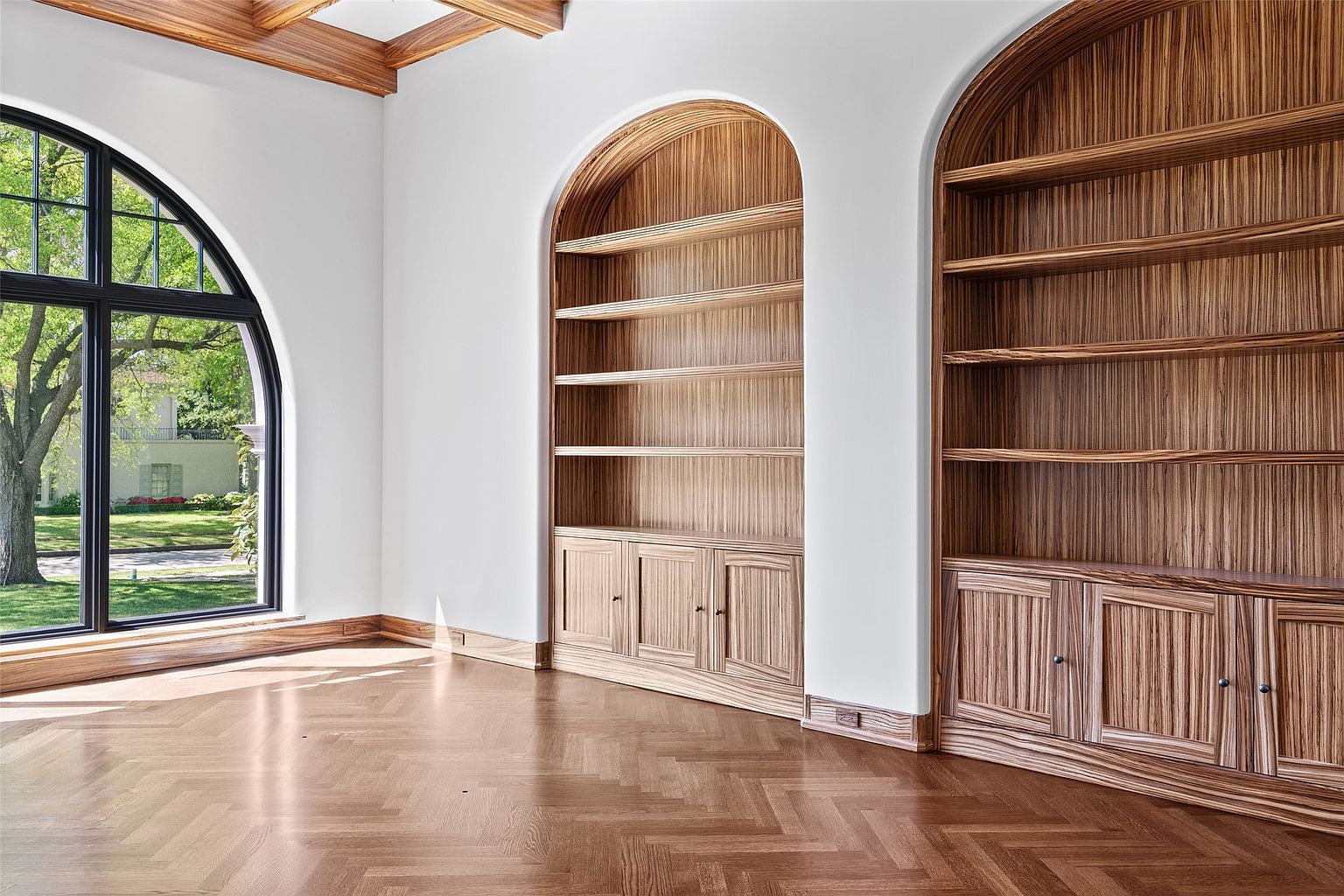 This interior shot showcases a sophisticated living room with custom built-in wooden bookshelves flanking a large arched window. The herringbone-patterned hardwood floors add warmth and texture, while the white walls provide a clean backdrop. The room exudes a sense of classic elegance and refined taste.