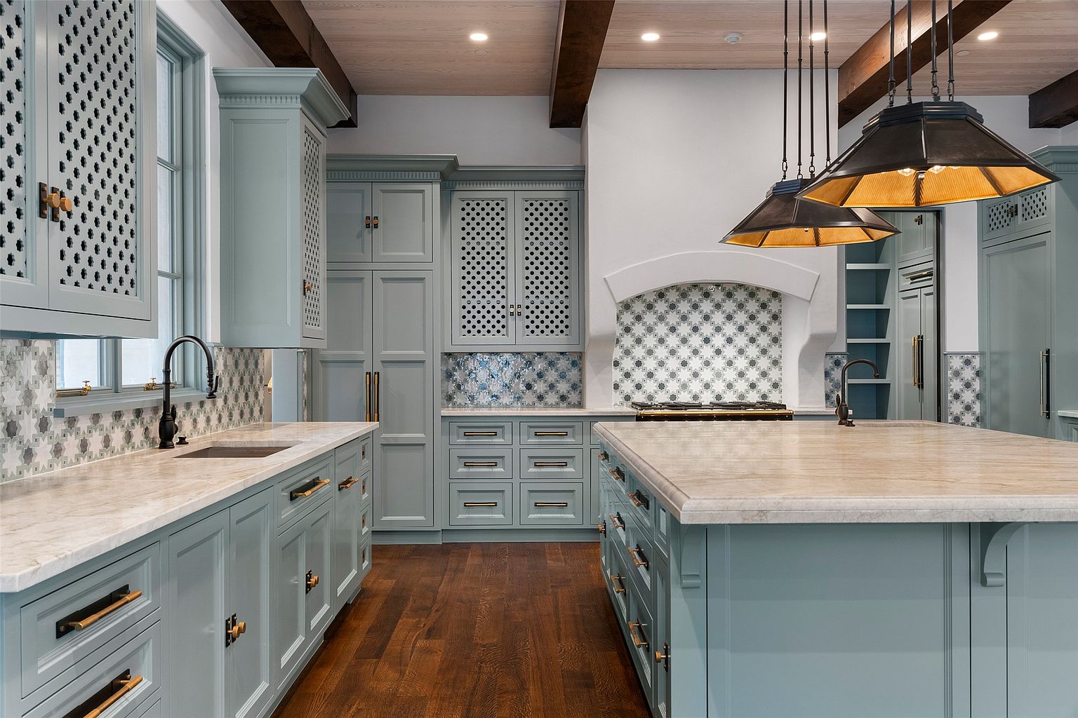 This is a beautifully designed kitchen featuring custom blue-grey cabinetry with unique latticework on some of the upper cabinets. The countertops are a light-colored stone, and the backsplash features a decorative tile pattern. A large island provides ample workspace, and pendant lights hang above it, adding a touch of elegance.