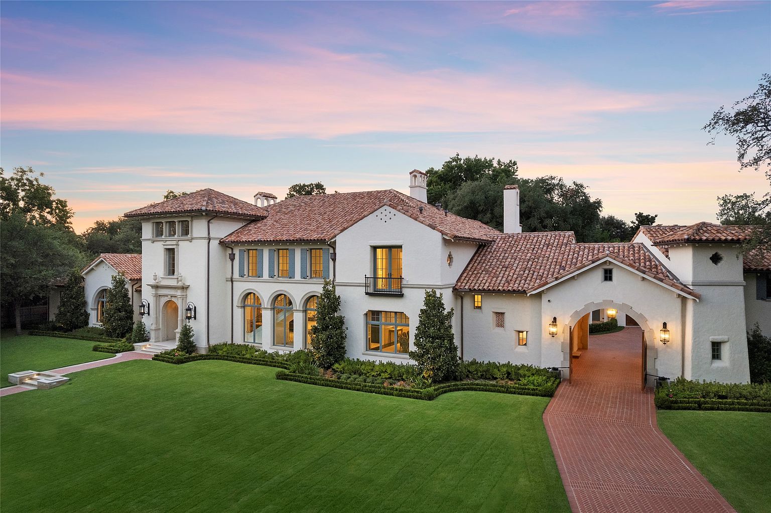 This is a stunning front view of a large, luxurious Mediterranean-style home. The house features a white stucco exterior, a red tile roof, and arched windows. A meticulously manicured green lawn and mature landscaping enhance the property's curb appeal, while a brick driveway leads to a grand arched entryway, creating an inviting and impressive approach.