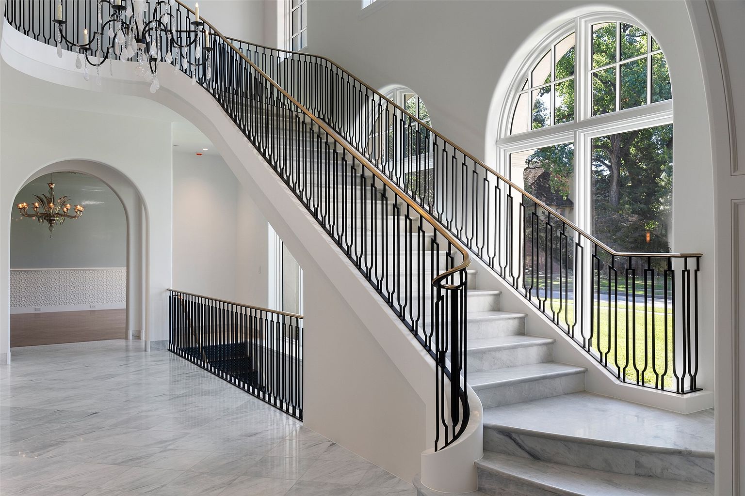 This interior shot showcases a grand staircase with marble steps and a modern black metal railing accented with a wooden handrail. Natural light floods the space through a large arched window, highlighting the pristine white walls and marble flooring. The overall impression is one of luxury and sophisticated design.