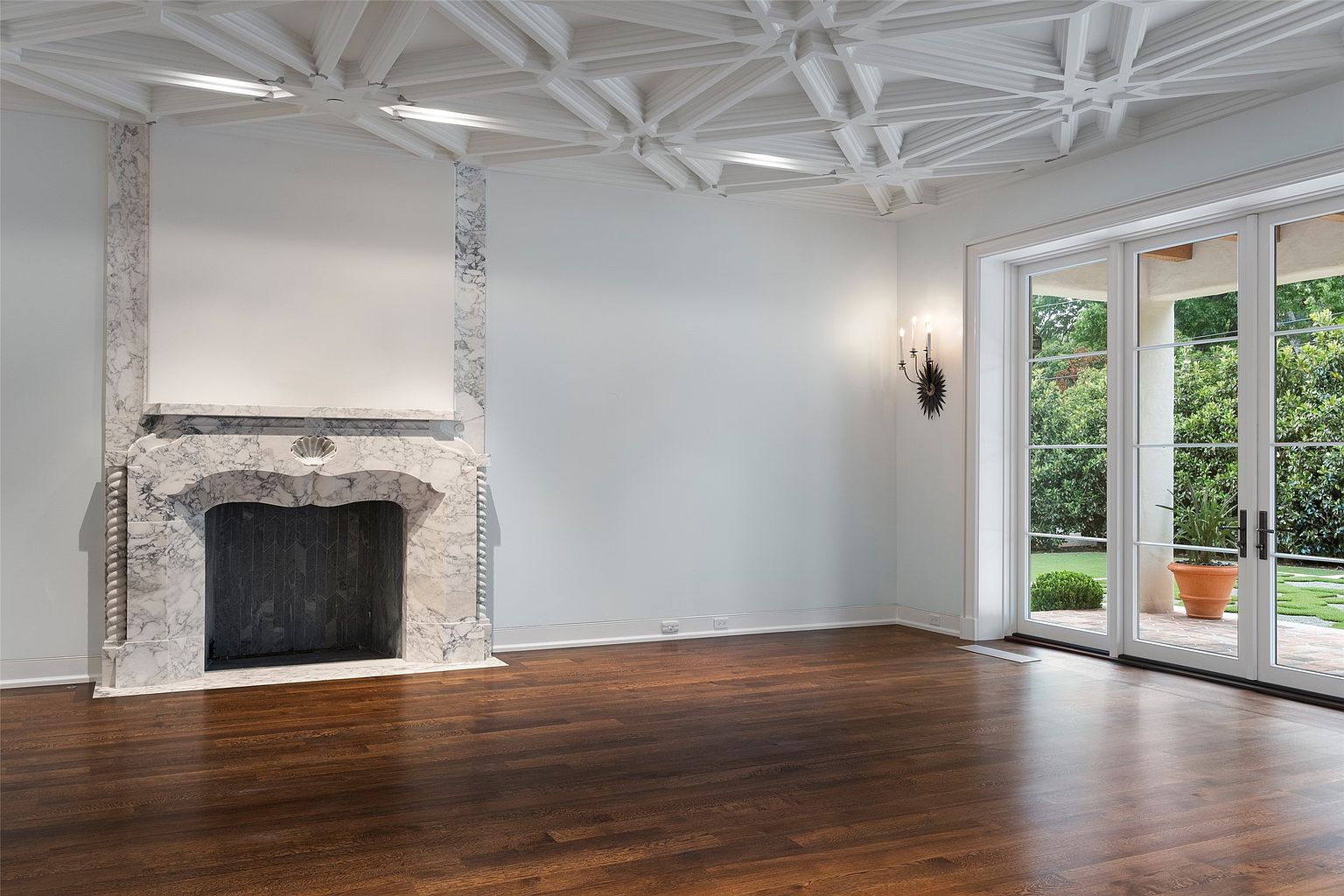 This is an interior shot of a living room featuring a marble fireplace, dark hardwood floors, and a decorative ceiling. Large glass doors lead to an outdoor area, providing natural light. The room is spacious and appears to be designed with a blend of classic and modern elements.