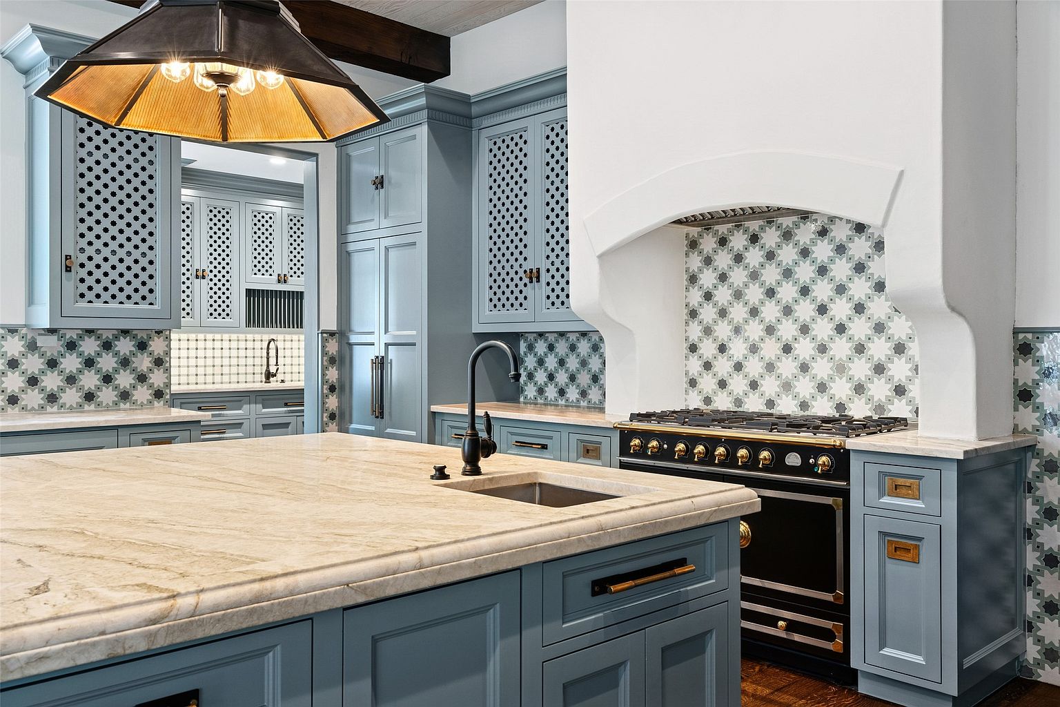 This is a beautifully designed kitchen featuring custom blue cabinetry with decorative perforated panels and a large island with a light-colored stone countertop. A black range with gold accents is positioned against a backsplash of patterned tiles, complemented by a white range hood. The kitchen is well-lit by a statement light fixture, creating a warm and inviting atmosphere.