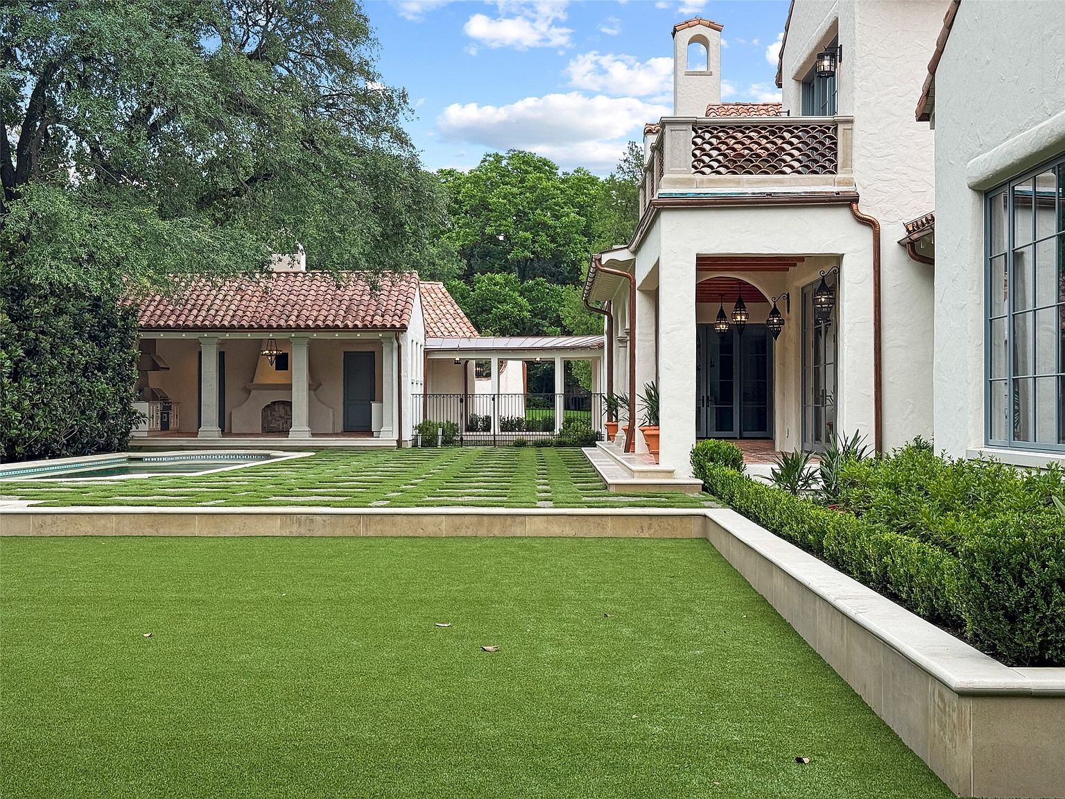 This image showcases a meticulously landscaped backyard featuring lush green grass, manicured hedges, and a stylish pool house with a terracotta tile roof. The property boasts a well-maintained lawn with geometric stone pathways, leading to a covered walkway and the main house, which exhibits elegant architectural details and a balcony. The overall impression is one of luxury and sophisticated outdoor living.
