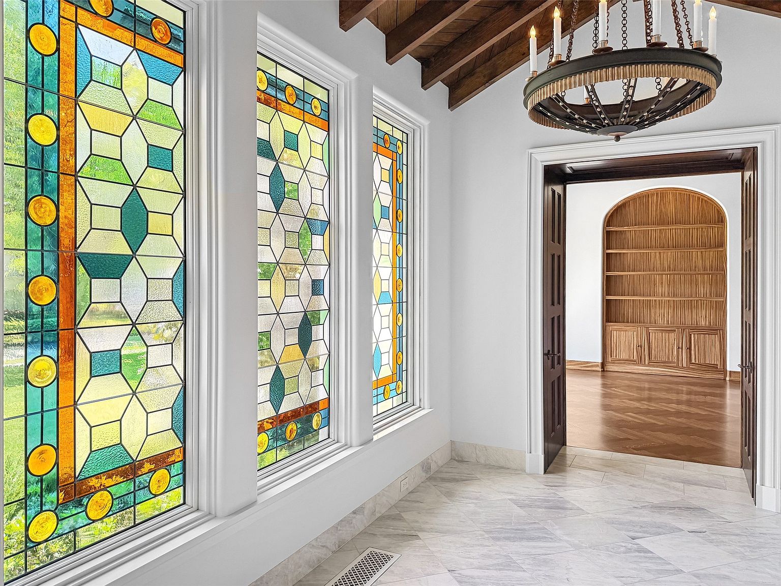This interior shot showcases a grand hallway with striking stained glass windows that flood the space with colorful light. The hallway features elegant marble flooring and leads to an open doorway revealing a wood-paneled room with built-in shelving. A decorative chandelier hangs from the ceiling, adding to the luxurious ambiance of the home.