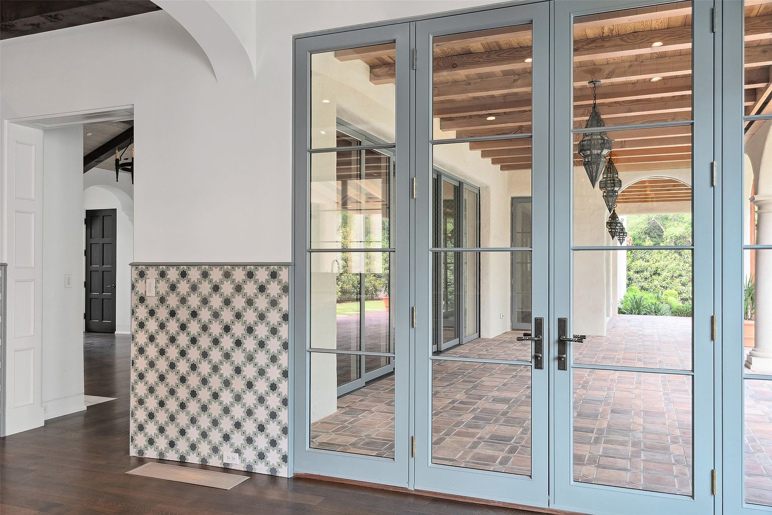 The image showcases an interior hallway featuring a decorative tiled wall and a set of elegant, light blue French doors leading to an outdoor patio. The dark hardwood flooring contrasts with the white walls and the light-colored doors, creating a sophisticated aesthetic. The view through the doors reveals a covered patio with brick flooring and a glimpse of lush greenery, enhancing the indoor-outdoor flow of the home.