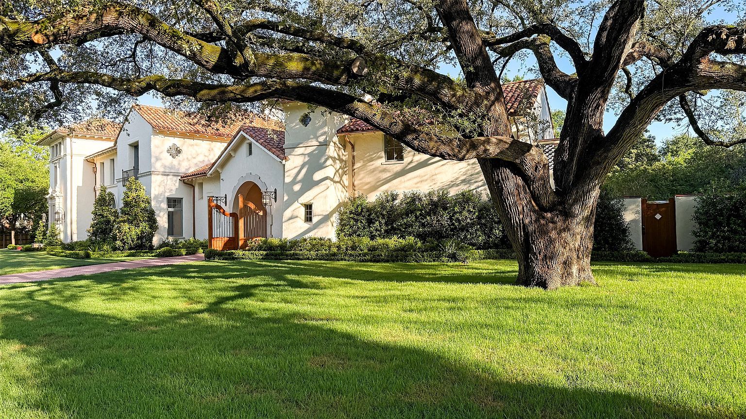 This image showcases the front exterior of a beautiful home with a Mediterranean-inspired design. The house features a light-colored stucco facade, a red tile roof, and an arched entryway with a wooden gate. A large, mature tree dominates the foreground, adding shade and character to the well-manicured lawn, enhancing the property's curb appeal.