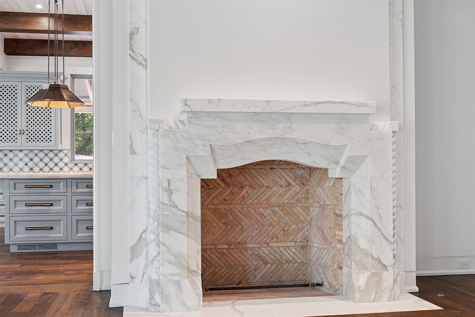 The image showcases a detailed view of a marble fireplace, highlighting its elegant design and craftsmanship. The fireplace features a unique herringbone brick pattern within the firebox, contrasting with the smooth marble surround. The perspective offers a glimpse into an adjacent kitchen area, adding depth and context to the architectural detail.