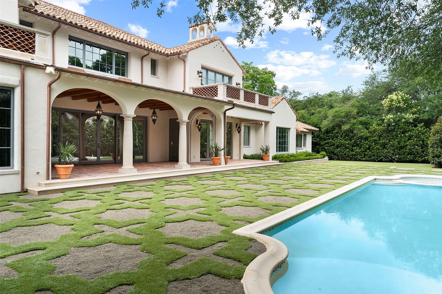This image showcases the rear exterior of a luxurious Mediterranean-style home, featuring a covered patio with arched columns, a tiled roof, and a balcony. The meticulously designed backyard includes a geometric grass and gravel pattern leading to a pristine swimming pool, all set against a backdrop of lush greenery, creating an inviting and elegant outdoor living space.