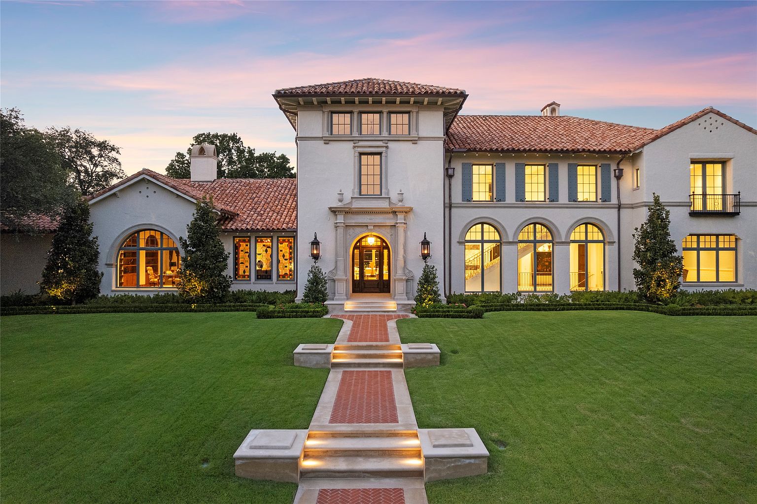This is a stunning front view of a large, luxurious home with a Mediterranean-inspired design. The house features a red tile roof, a prominent central tower, and arched windows that add to its elegant appeal. A meticulously manicured lawn and a brick pathway lead to the grand entrance, creating a welcoming and impressive approach.