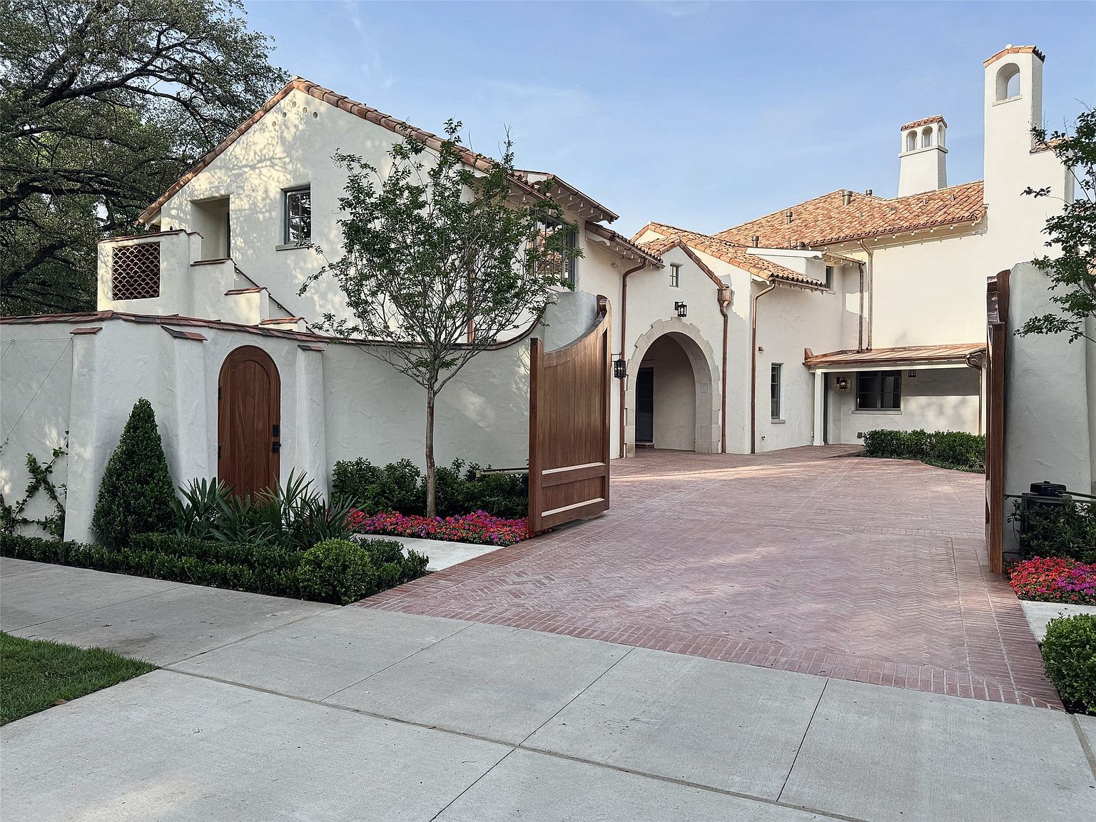 This is a front view of a luxurious, Mediterranean-style home. The house features a white stucco exterior, a red tile roof, and a brick driveway. A wooden gate provides access to the property, and lush landscaping adds to the curb appeal. The overall impression is one of elegance and privacy.