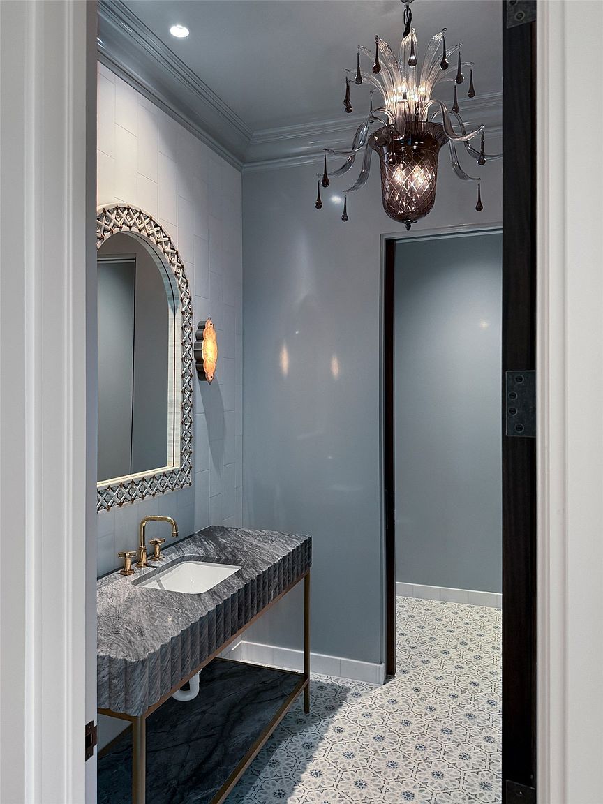 This is a stylish guest bathroom featuring a unique vanity with a gray marble countertop and a decorative mirror. The walls are painted a soft gray, and the floor is tiled with a patterned design. A decorative chandelier hangs from the ceiling, adding a touch of elegance to the space.