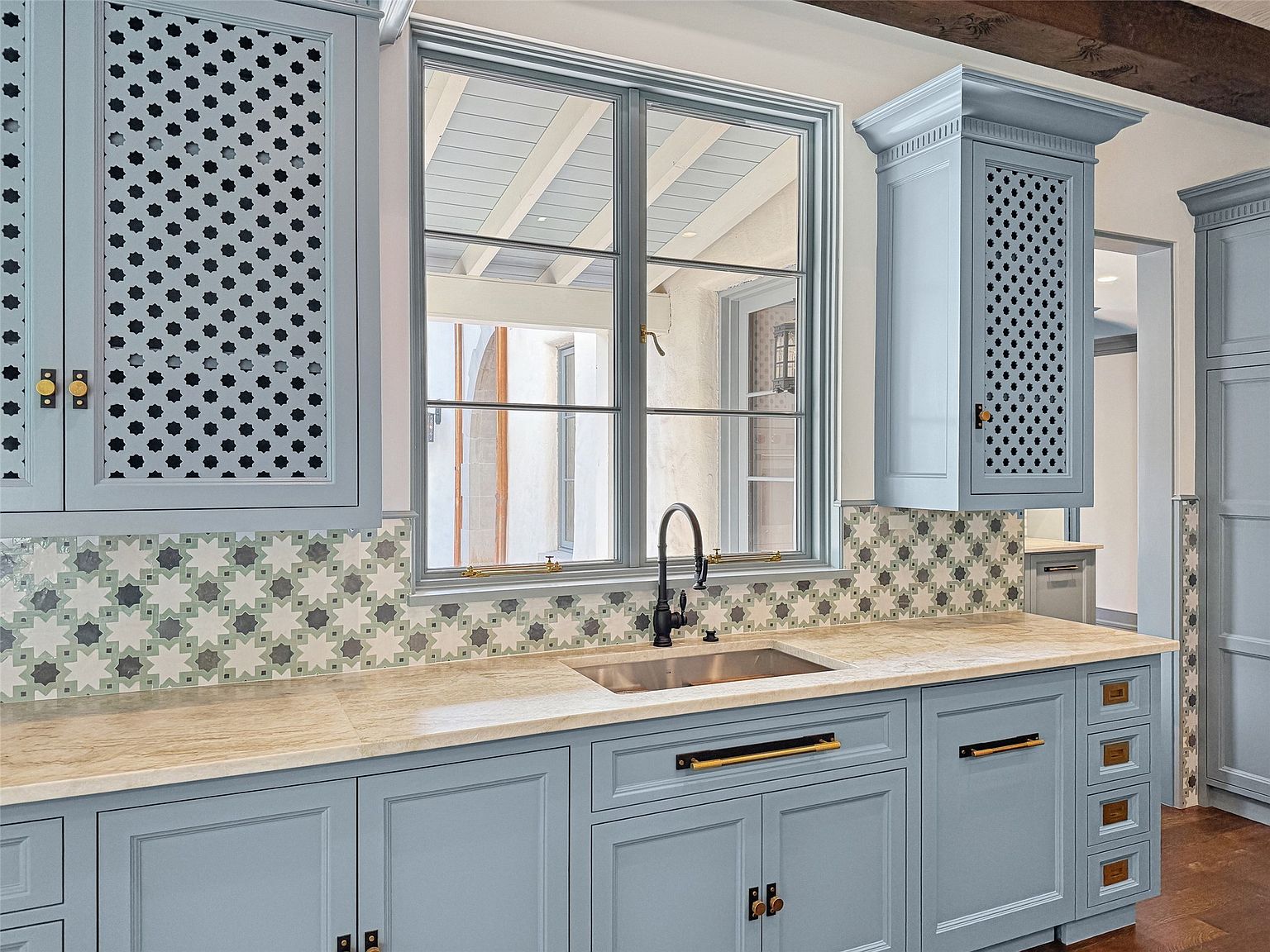This is a well-lit kitchen featuring custom blue cabinetry with decorative perforated panels and brass hardware. A large window sits above the sink, providing natural light and a view to the outside. The countertops are a light-colored stone, and the backsplash features patterned tiles, adding a touch of elegance to the space.