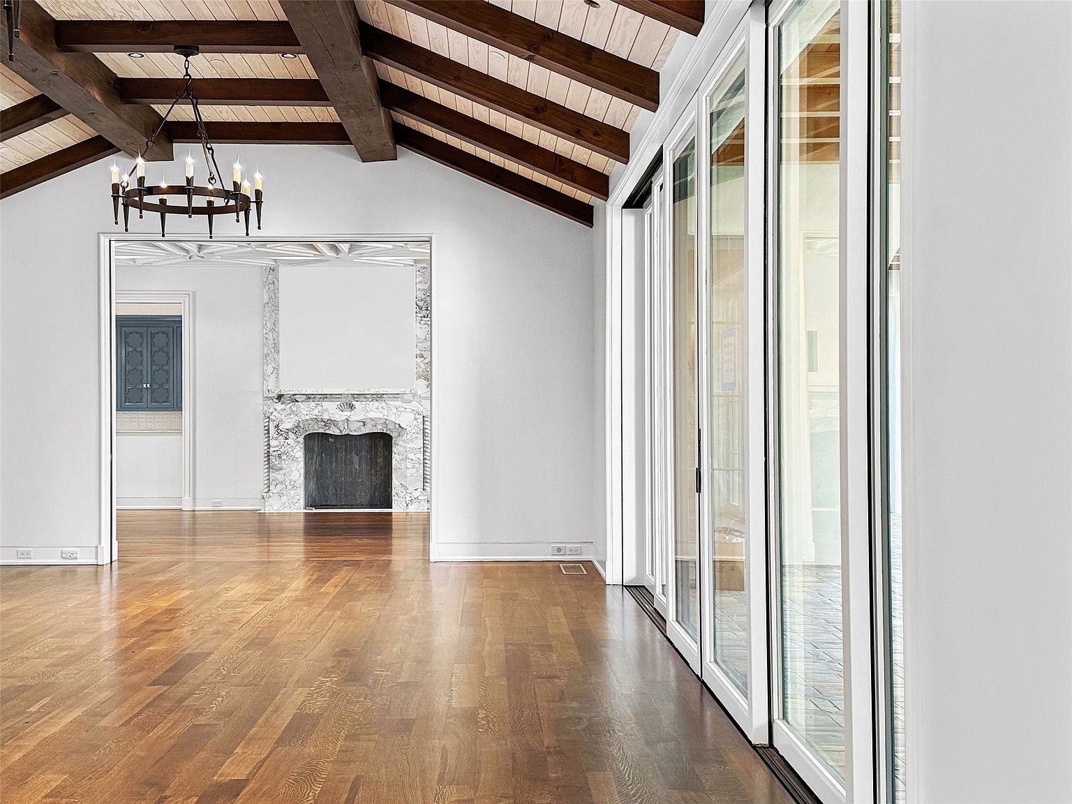 This interior shot showcases a spacious living room with hardwood floors, a vaulted ceiling with exposed beams, and a decorative chandelier. A marble fireplace is visible through an archway, and large sliding glass doors provide ample natural light. The room exudes a sense of elegance and openness, perfect for entertaining or relaxing.