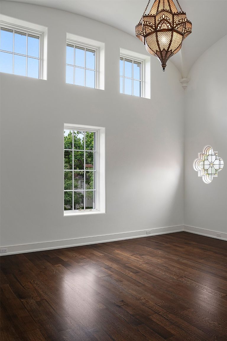 This is an interior shot of a living room featuring dark hardwood floors and white walls. The room has multiple windows, including three smaller ones near the ceiling and a larger one providing a view of greenery outside. An ornate chandelier hangs from the ceiling, and a decorative window is visible on the right wall, creating a bright and airy atmosphere.