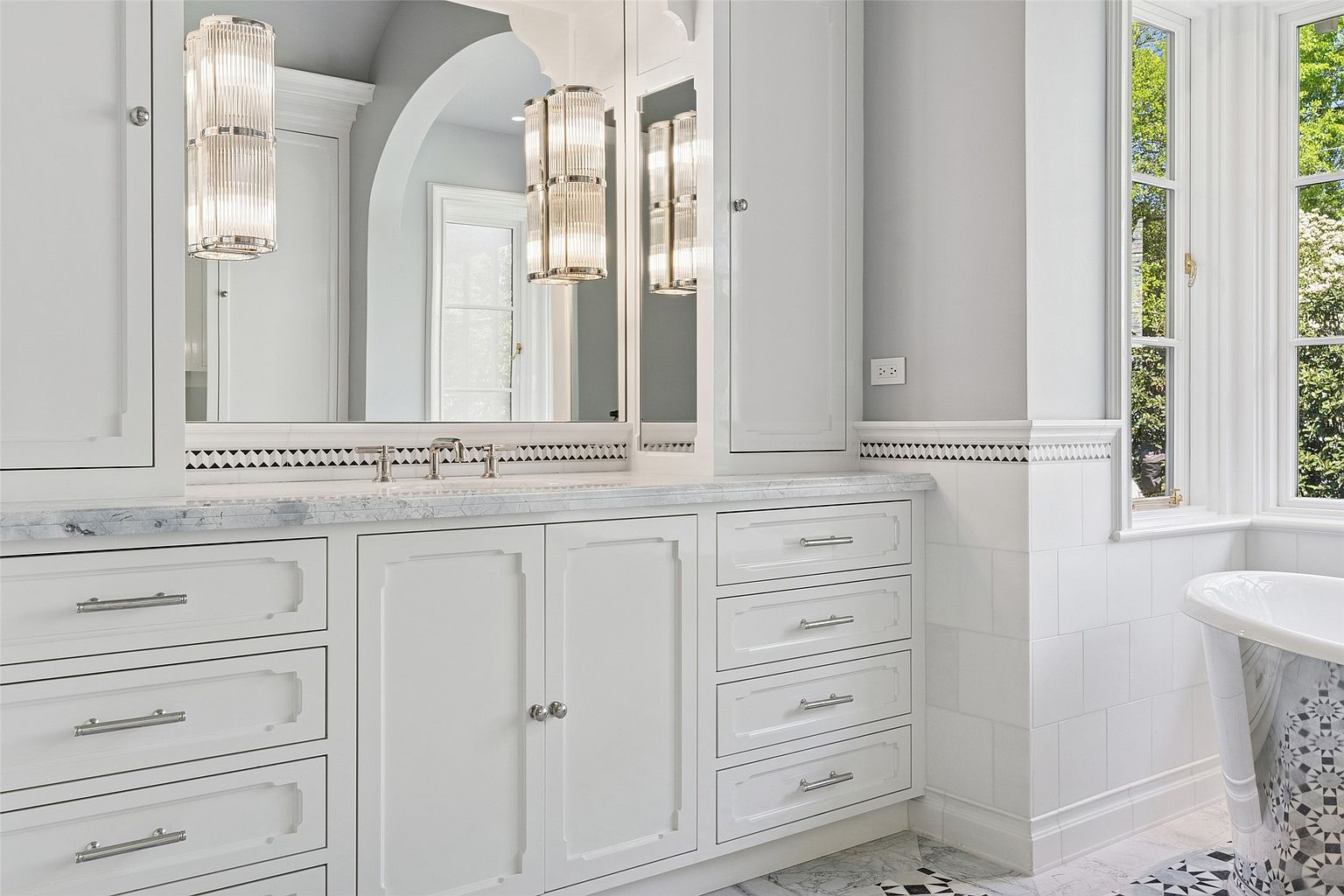 This is a well-lit primary bathroom featuring a white vanity with marble countertops and silver hardware. The vanity has both drawers and cabinets, and the room is decorated with black and white patterned floor tiles and a matching bathtub. The overall impression is clean, elegant, and luxurious.