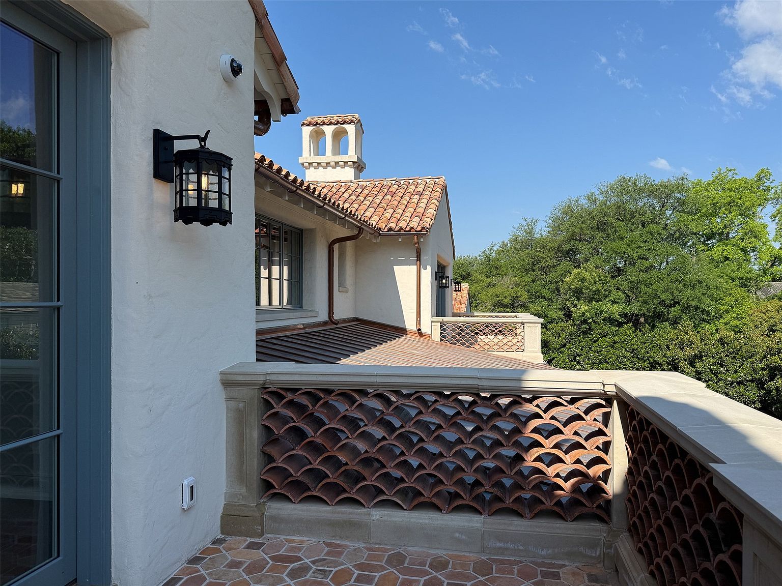 This image showcases a charming balcony with a unique architectural design. The balcony features a decorative terracotta tile screen, a tiled floor, and a stone railing, creating a Mediterranean-inspired aesthetic. The view includes a glimpse of the surrounding landscape and the building's architectural details, such as the tiled roof and a small tower.
