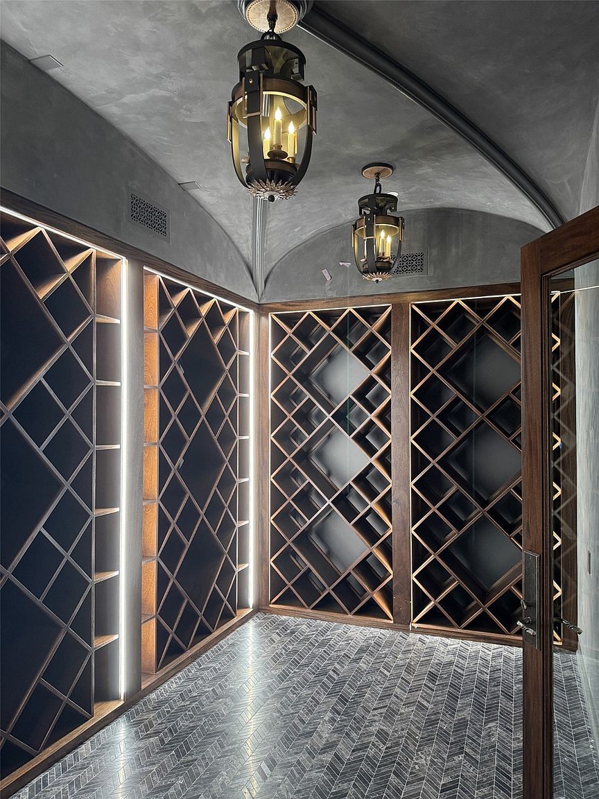 This is an interior shot of a wine cellar featuring custom wooden wine racks with diamond-shaped compartments, illuminated by vertical LED strips. The floor is tiled in a herringbone pattern, and the ceiling is arched and finished in a dark, textured material. Two ornate pendant lights hang from the ceiling, and a partially open wooden door is visible on the right, suggesting a sophisticated and well-designed space for wine storage.