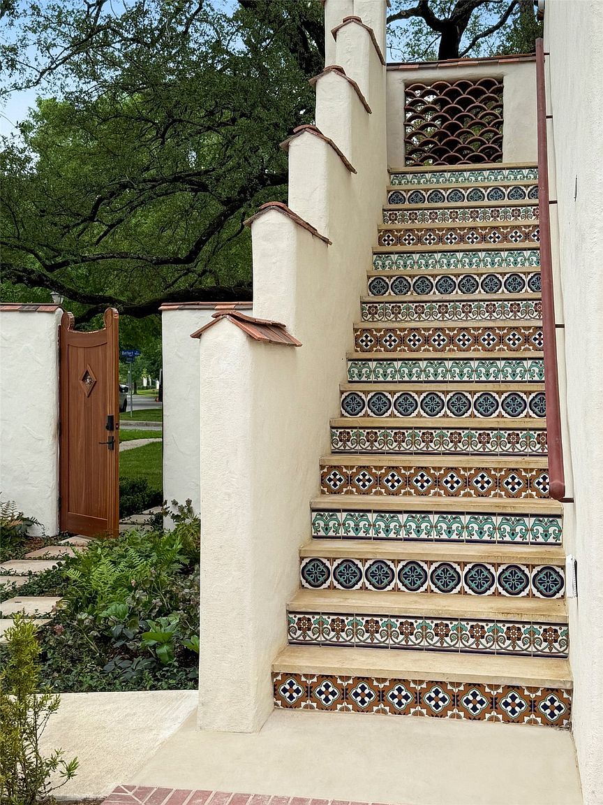 This image showcases an inviting entryway with decorative tiled stairs leading up to a building. The stairs are adorned with colorful, patterned tiles, adding a unique and artistic touch. A wooden gate and lush greenery complement the architectural details, creating a warm and welcoming atmosphere.