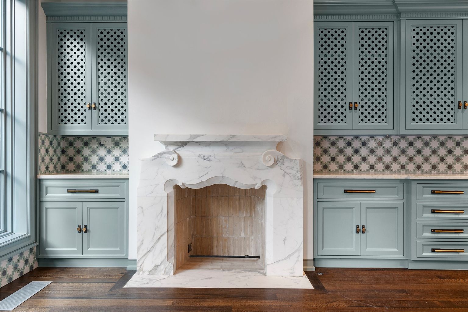 This interior shot showcases a sophisticated living room with custom built-in cabinetry flanking a marble fireplace. The cabinets are a muted blue-green color with decorative latticework on the upper doors and stylish hardware. The fireplace is the focal point, featuring elegant marble detailing and a clean, modern design.