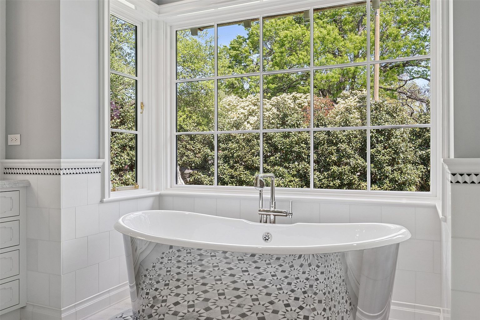 This is a bright and elegant primary bathroom featuring a freestanding bathtub with a decorative patterned exterior. The room is well-lit by a large window offering a view of lush greenery. White tilework and classic fixtures contribute to a clean and luxurious aesthetic.