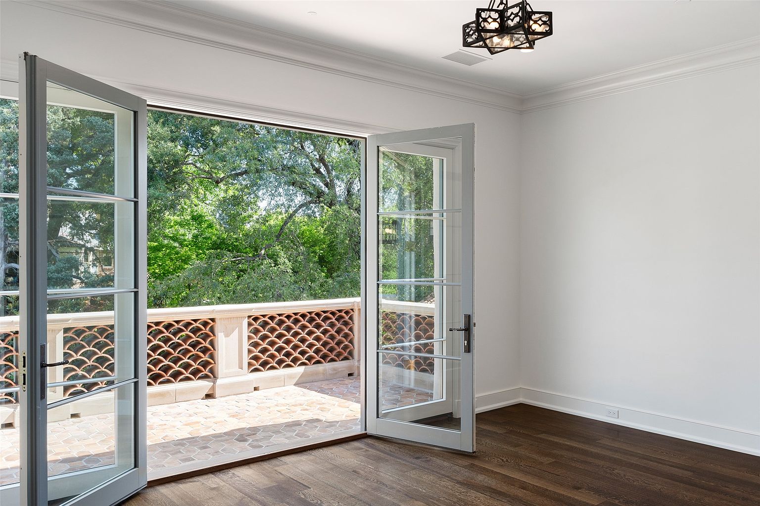 This interior shot showcases a bright bedroom with an open balcony. The room features hardwood flooring, white walls, and a decorative ceiling light fixture. French doors lead to a balcony with a patterned terracotta railing, offering a view of lush greenery.
