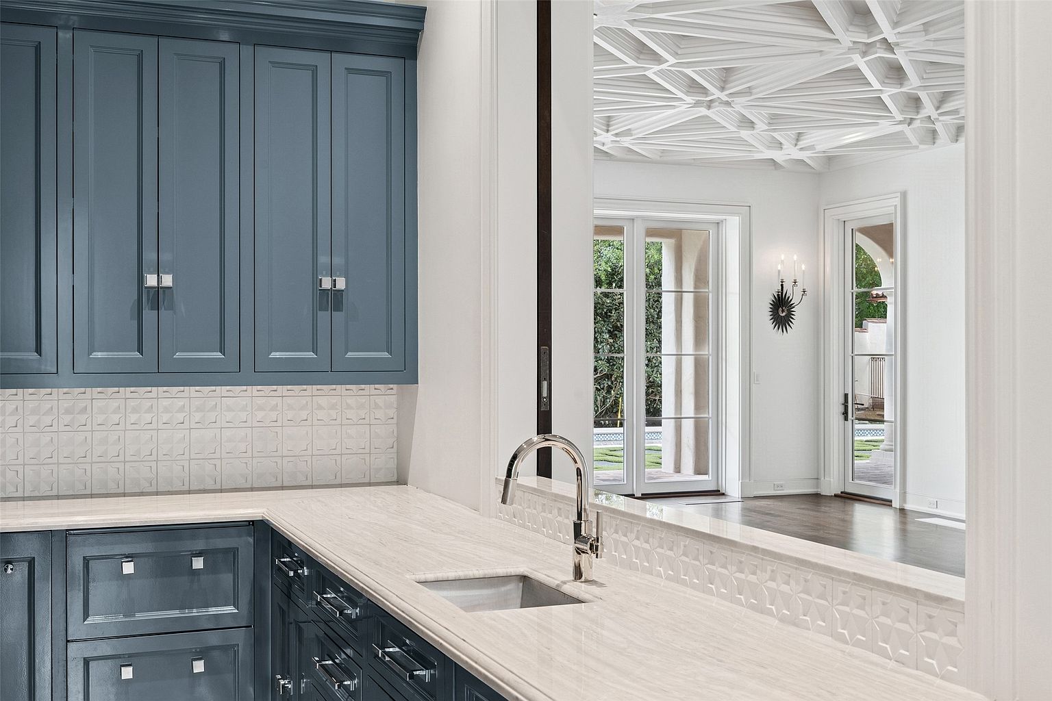 This is a detailed interior shot of a kitchen featuring custom blue cabinetry with modern hardware, light-colored countertops, and a stainless steel sink and faucet. The backsplash is a textured white tile, and the kitchen opens into a bright, white room with a decorative ceiling and access to an outdoor area through French doors. The perspective is from within the kitchen, looking out towards the adjacent room, creating a sense of openness and connection.