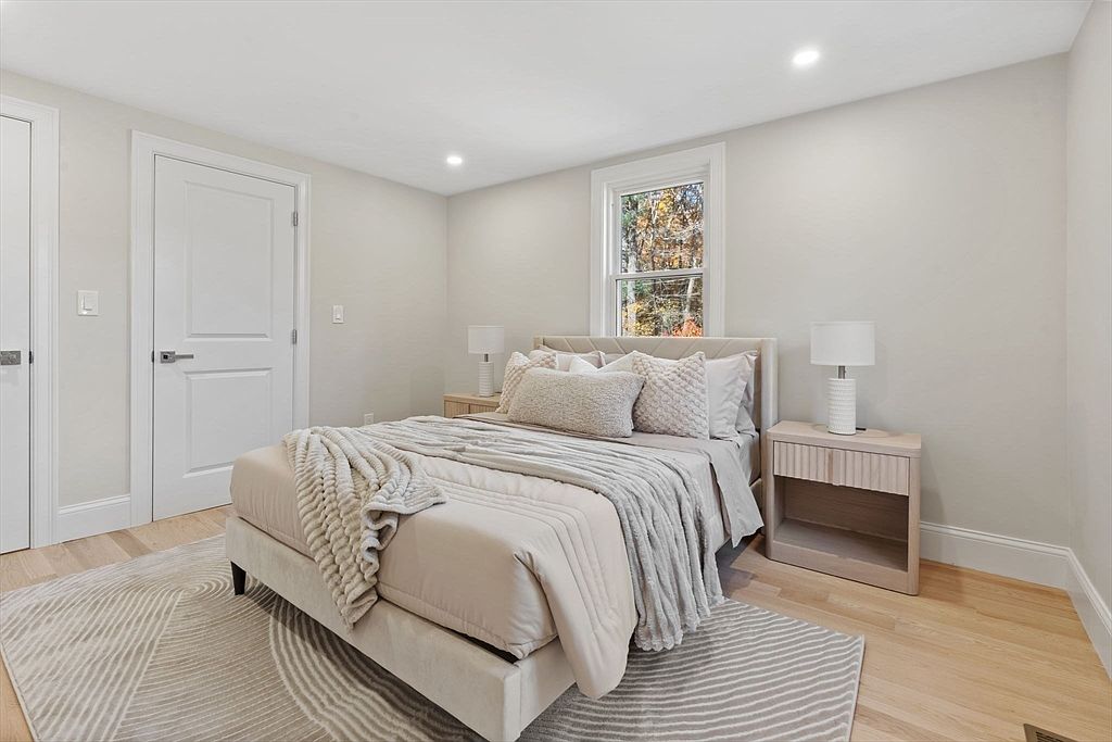 This is a bright and airy primary bedroom featuring a neutral color palette. The room includes a bed with a padded headboard, two matching nightstands with lamps, and a stylish area rug. A window provides natural light, enhancing the serene and inviting atmosphere of the space.