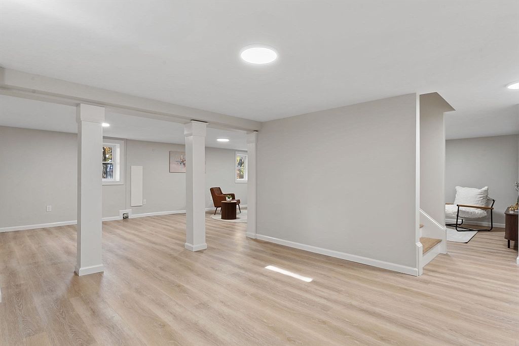 This is a spacious, finished basement featuring light wood-look flooring and neutral gray walls. The room is well-lit with recessed lighting and natural light from windows. Structural support columns are integrated into the design, and there's a glimpse of a seating area with a chair and side table in the background, suggesting a comfortable and versatile living space.
