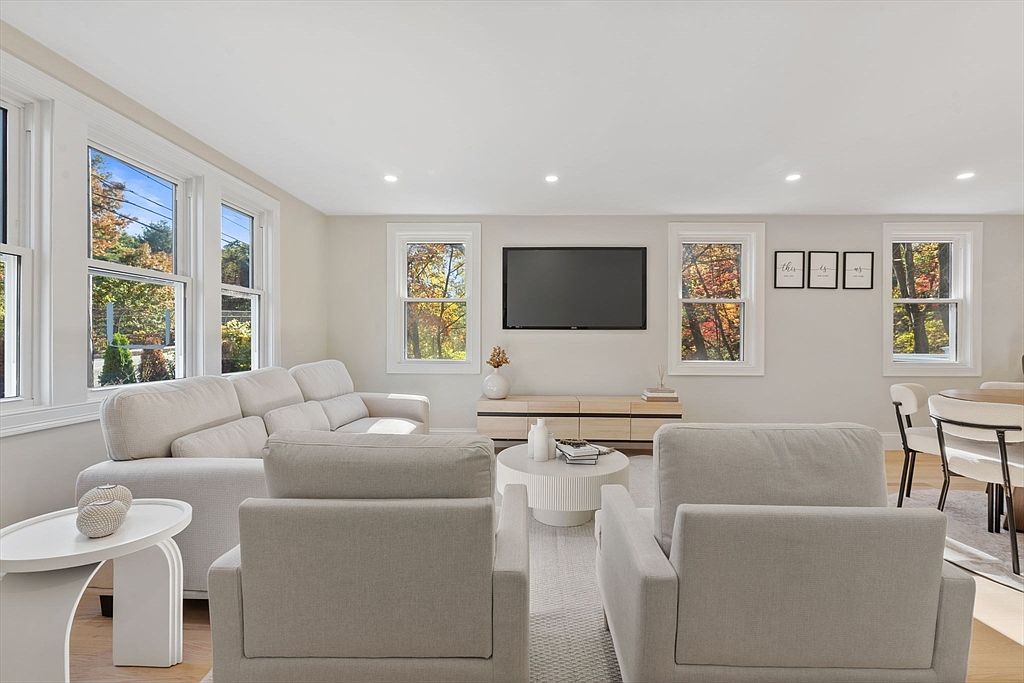 This is a bright and airy living room featuring neutral tones and modern furnishings. The room includes a comfortable sofa, two armchairs, a round coffee table, and a media console with a large flat-screen TV. Large windows provide ample natural light and views of the outdoors, creating a welcoming and serene atmosphere.