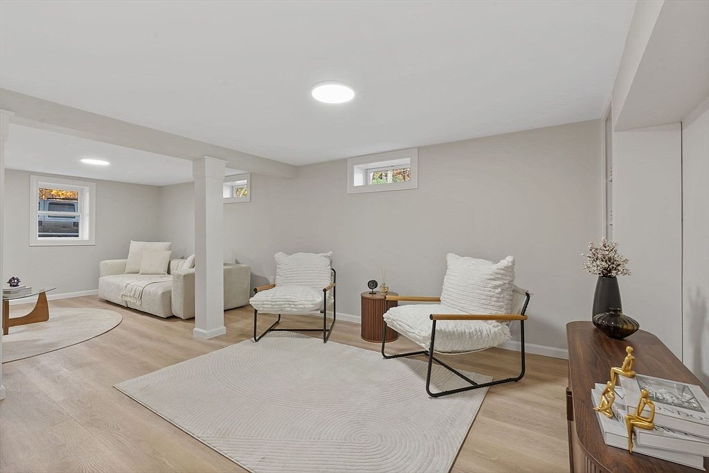 This is a bright and airy living room featuring modern furniture and neutral tones. The room includes a light-colored sofa, two armchairs with white cushions, a wooden side table, and a console table with decorative items. The flooring is light wood, and a large area rug anchors the seating arrangement, creating a cozy and inviting atmosphere.