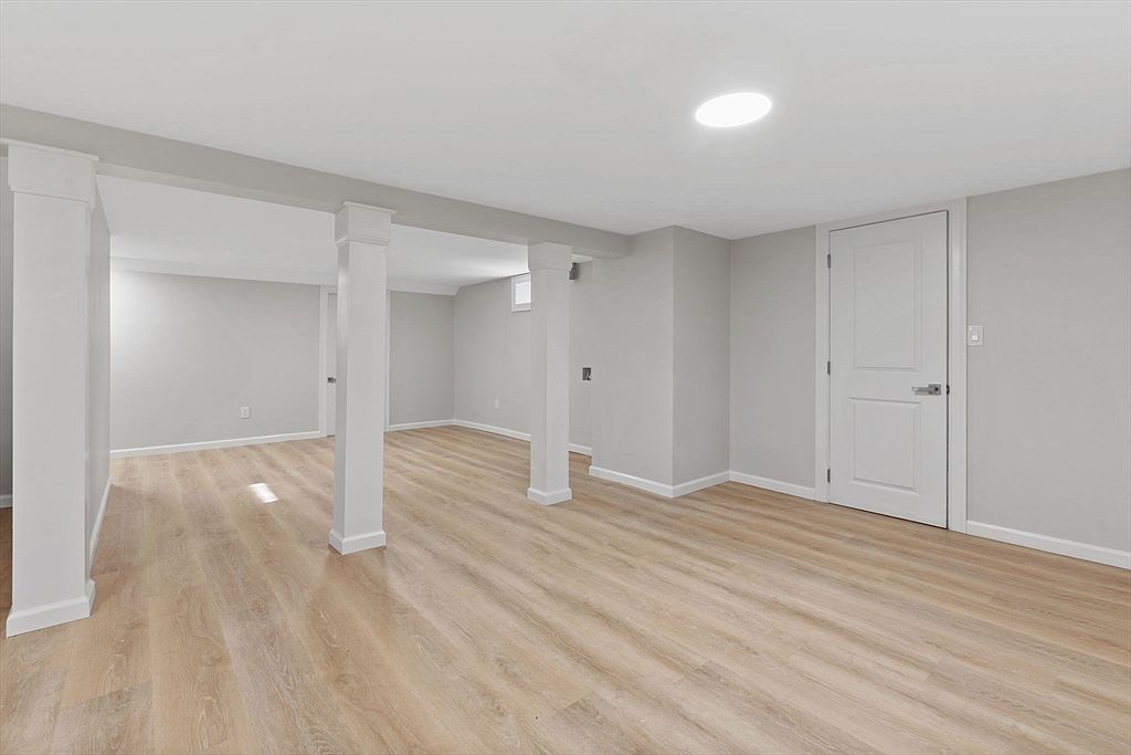 The image showcases a finished basement with light wood-look flooring and light gray walls. White support columns and trim provide a clean, modern aesthetic. A white door is visible on the right, suggesting access to another area or storage.