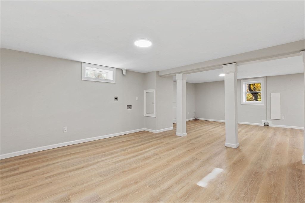 The image showcases a finished basement with light wood-look flooring and light gray walls. White support columns and beams are visible, adding architectural interest. A small window provides natural light, and the overall impression is clean and modern, suggesting a versatile space for recreation or storage.
