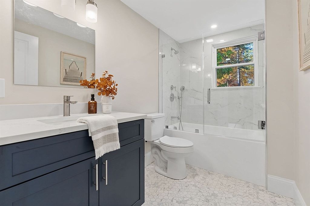 This is a well-lit bathroom featuring a navy blue vanity with a white countertop, a modern faucet, and a rectangular mirror. A white toilet sits beside the vanity, and a glass-enclosed bathtub/shower combination is visible in the background, showcasing marble-like tiling. The floor is covered in a light-colored, patterned tile, creating a clean and stylish space.