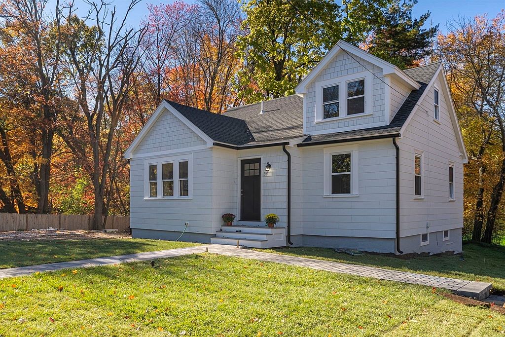 This is a charming front view of a newly renovated home. The house features white siding, a dark front door, and well-maintained landscaping with a stone walkway leading to the entrance. The surrounding trees showcase autumn colors, adding to the property's curb appeal.