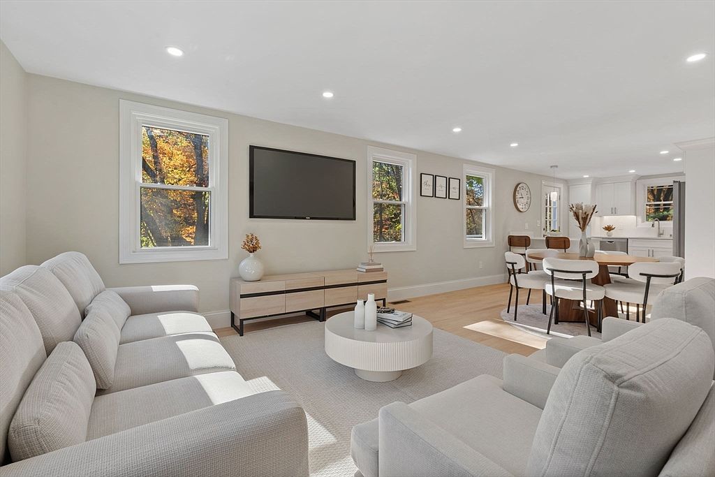 This is a bright and airy living room featuring a large sectional sofa, a modern coffee table, and a media console beneath a flat-screen TV. Natural light floods the space through multiple windows, highlighting the neutral color palette and creating a welcoming atmosphere. The open floor plan seamlessly connects the living area to the dining space and kitchen.
