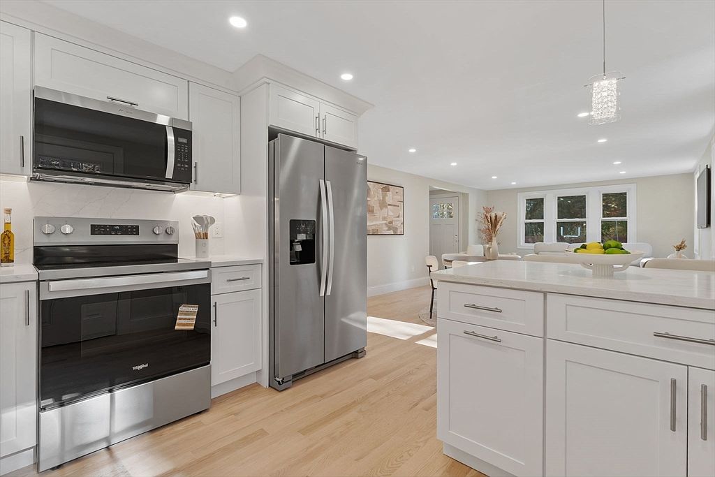 This is a bright and modern kitchen featuring white cabinets, stainless steel appliances, and light hardwood floors. The kitchen is open to the dining area, creating a spacious and inviting atmosphere. The stainless steel refrigerator and oven are prominent features, complemented by the white countertops and cabinetry.