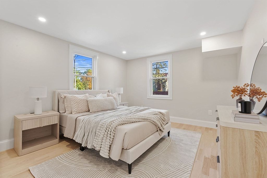 This is a bright and airy primary bedroom featuring neutral tones and natural light. The room is furnished with a bed, side table, dresser, and a decorative rug. The overall impression is clean, comfortable, and inviting, perfect for relaxation.