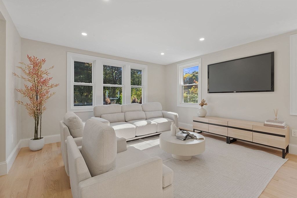 This is a bright and airy living room featuring a neutral color palette. The room is furnished with a light-colored sofa, armchairs, a round coffee table, and a media console beneath a large television. Natural light floods the space through multiple windows, creating a welcoming and comfortable atmosphere.