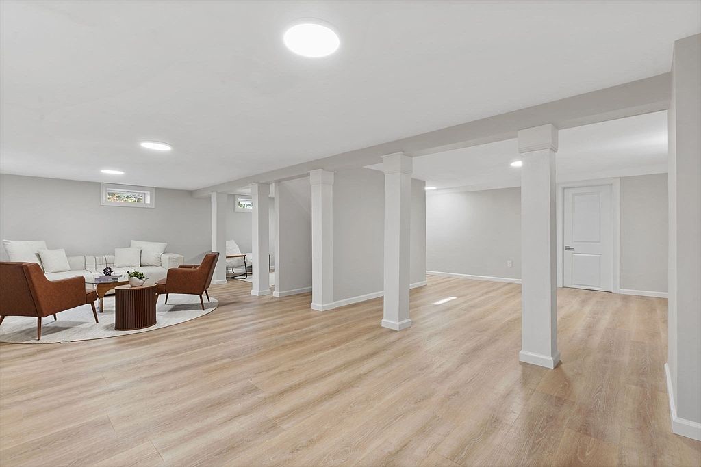 The image showcases a spacious, finished basement with light wood-look flooring and white walls. Architectural columns provide support and visual interest, while a seating area with modern furniture adds a touch of comfort. The room is well-lit with recessed lighting, creating a bright and inviting atmosphere.