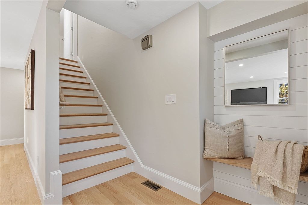 This interior shot showcases a hallway with a staircase leading to an upper level. The stairs feature wooden treads and white risers, complemented by light hardwood flooring. A built-in bench with a pillow and throw adds a touch of comfort and style to the space, while a mirror reflects light and creates a sense of openness.