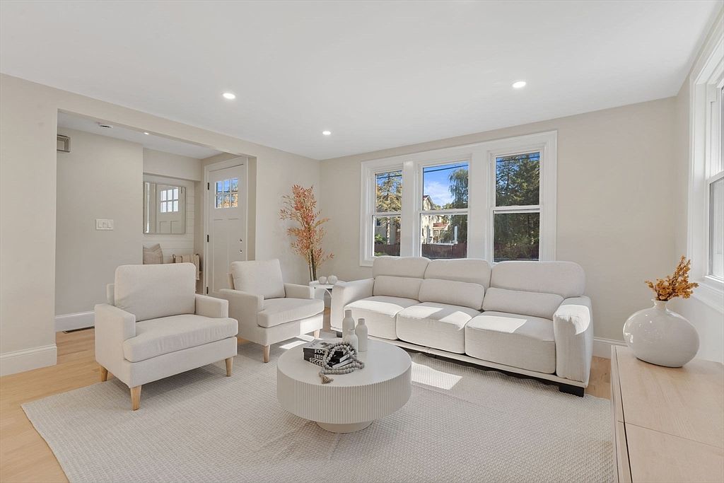 The image showcases a bright and inviting living room with a neutral color palette. The room features a white sofa, two armchairs, a round coffee table, and a light-colored rug. Natural light floods the space through a large window, creating a warm and welcoming atmosphere.