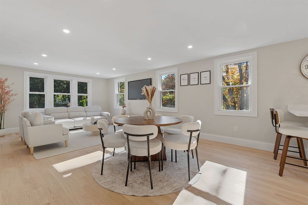 This is an interior shot of a living and dining area. The living room features a white sectional sofa and a light-colored rug, while the dining area has a round wooden table with six chairs. Large windows provide natural light, and the overall aesthetic is bright and modern, creating a welcoming and spacious atmosphere.